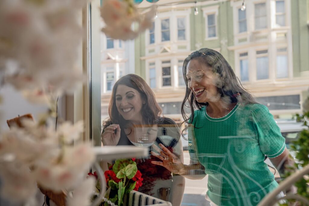 Two women smile and look through a shop window display decorated with flowers and various items, with a city street visible behind them.