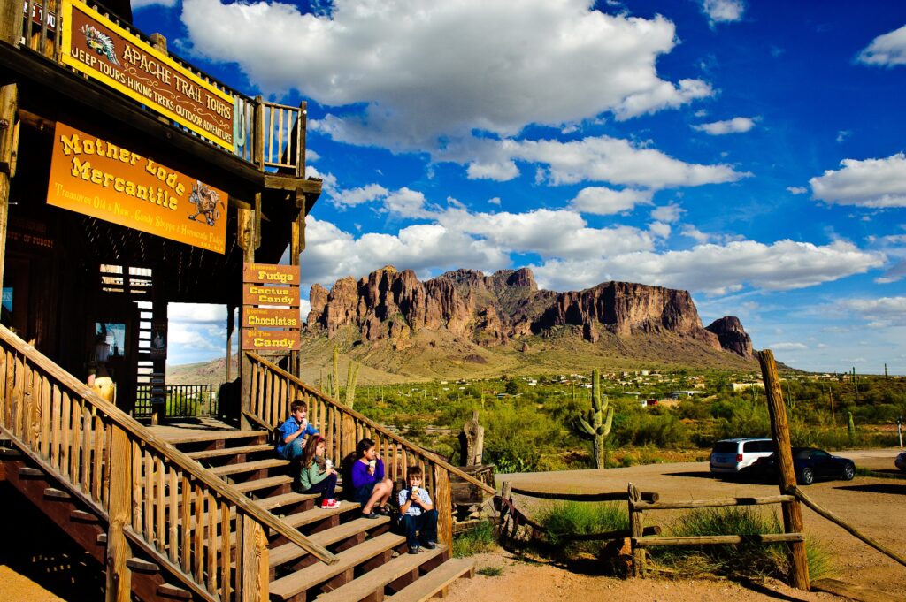 People sit on wooden steps outside a rustic store with a yellow sign, with desert landscape and rocky mountains under a partly cloudy sky in the background.