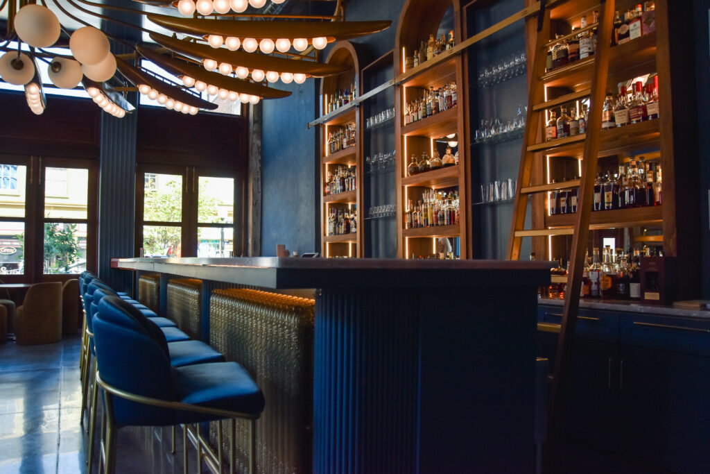 A modern bar with blue stools, a marble counter, shelves filled with bottles and glassware, a wooden ladder, and large round ceiling lights.