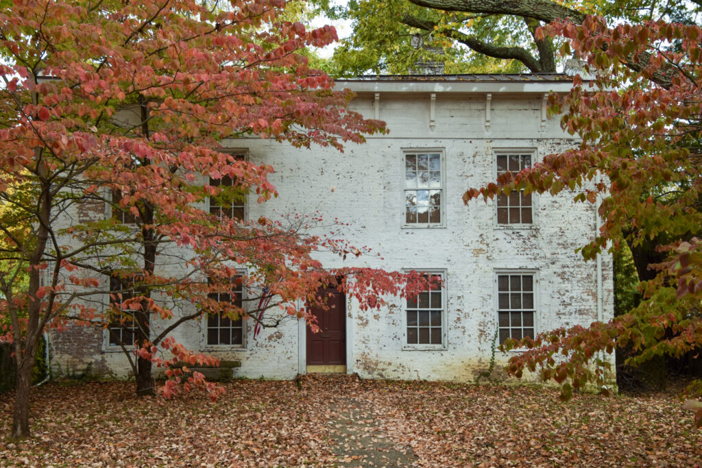 Two-story white brick house with a red front door, surrounded by trees with red and orange autumn leaves. Fallen leaves are scattered on the ground in front of the house.
