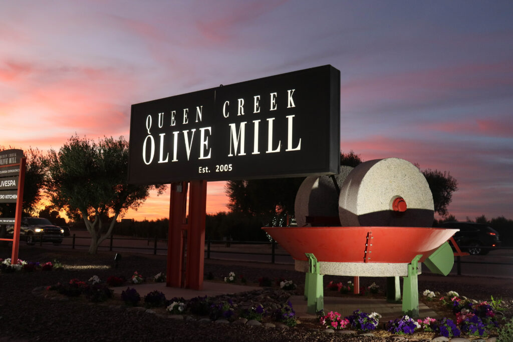 A lit sign reads “Queen Creek Olive Mill Est. 2005” beside a large olive press with trees and flowers in the background at sunset.