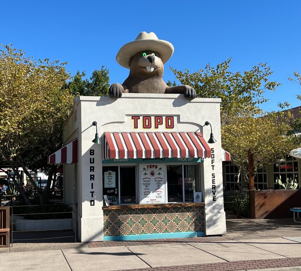 Small building with a red and white striped awning, sign reading “TOPO,” and a large animal statue in a cowboy hat on the roof. Trees and benches surround the structure under a clear blue sky.