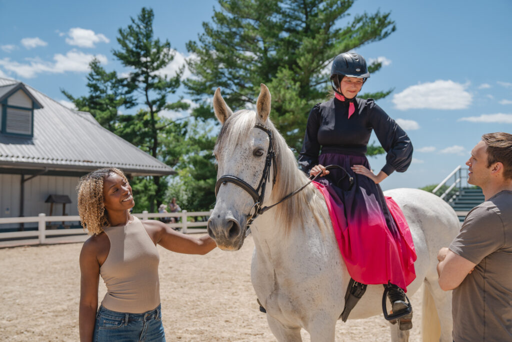 A woman in a helmet and dress sits on a white horse, while another woman pets the horse and a man stands nearby. They are outside by a building and trees on a sunny day.
