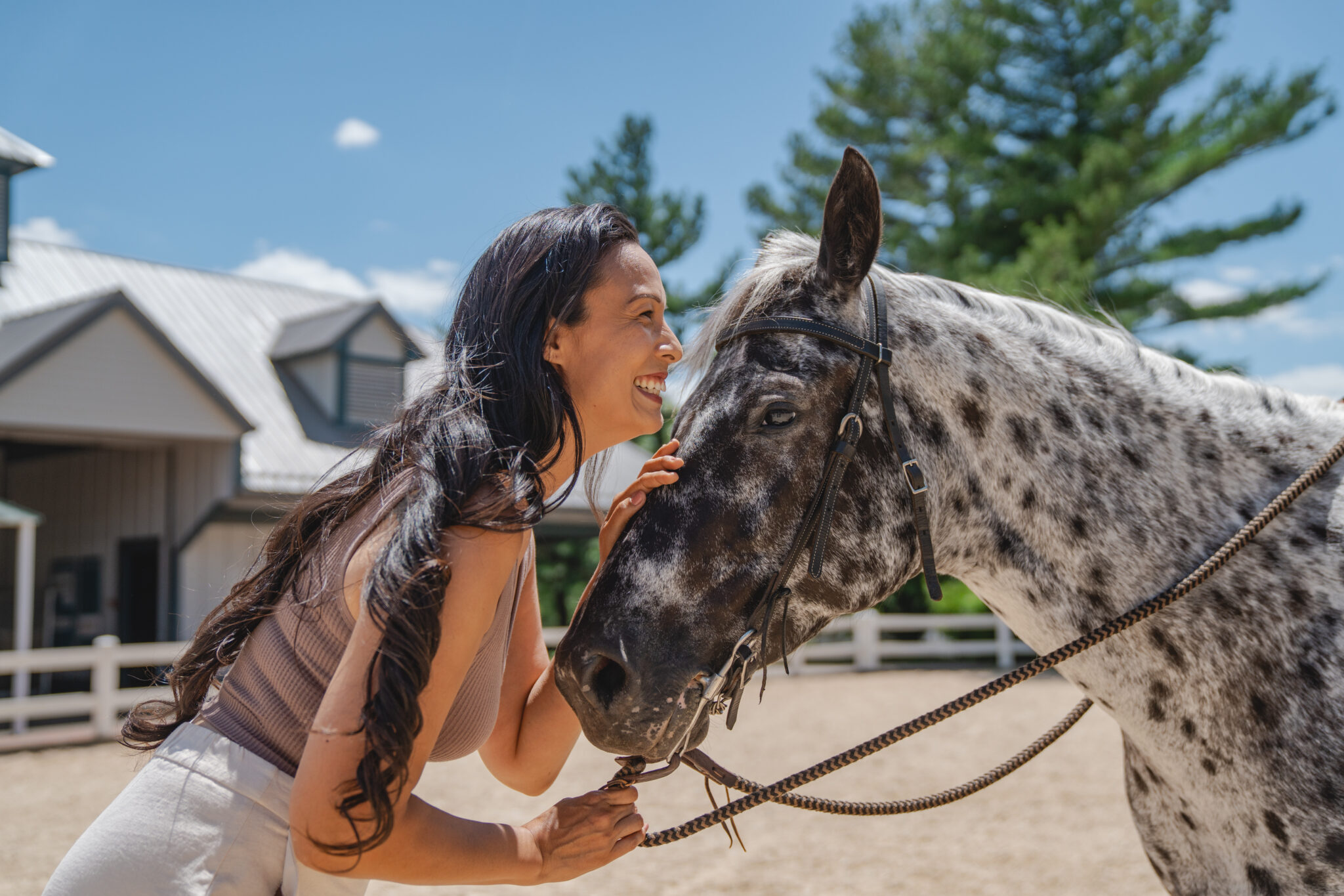 Woman smiles and leans affectionately against the head of a black and white spotted horse outdoors near a stable.