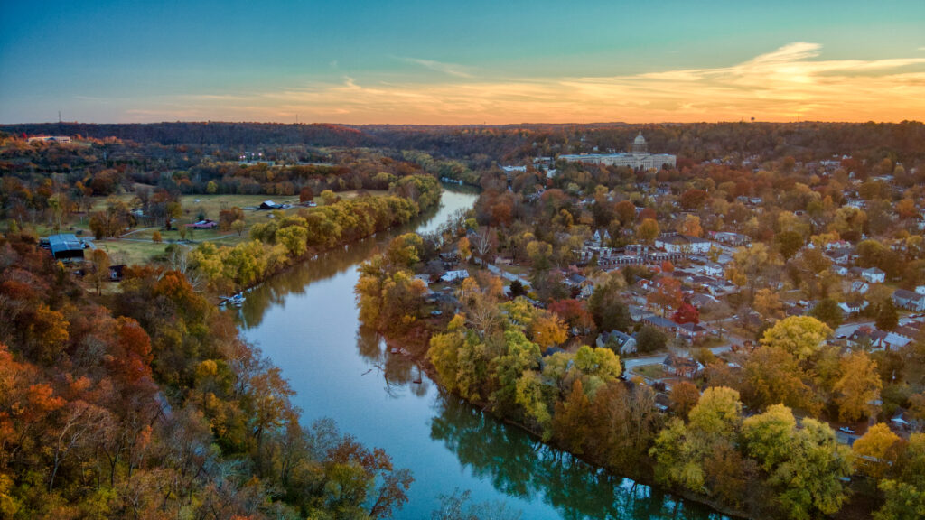 A river winds through a tree-filled landscape with houses and buildings nearby, seen from above at sunset.