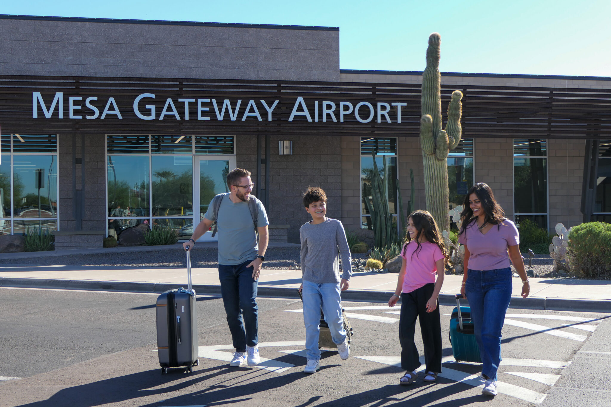 A family of four with luggage walks outside Mesa Gateway Airport on a sunny day, with desert plants and a large cactus visible nearby.
