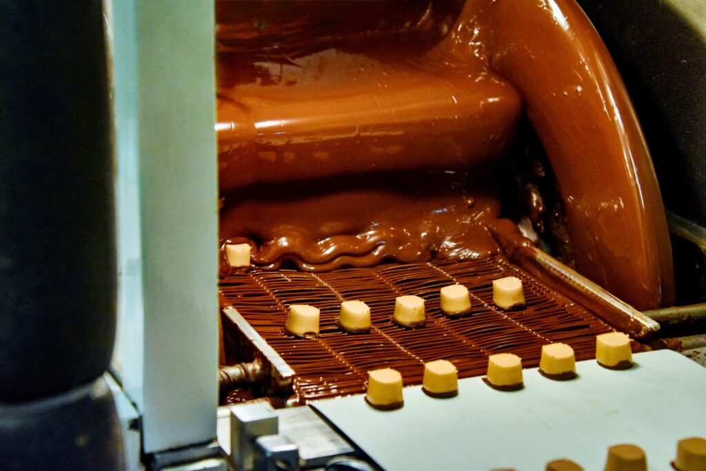 Confectionery factory machine coating small pieces of candy or biscuits with a layer of liquid chocolate on a conveyor belt.