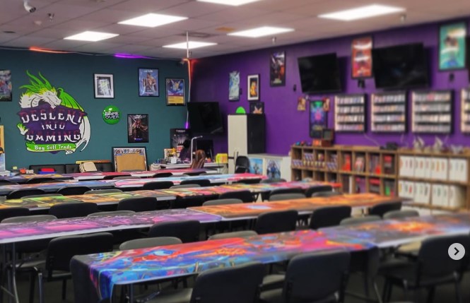 A game store with rows of tables covered in colorful playmats, chairs, wall posters, TV screens, shelves of card boxes, and a large mural reading “Descent Into Gaming.”.