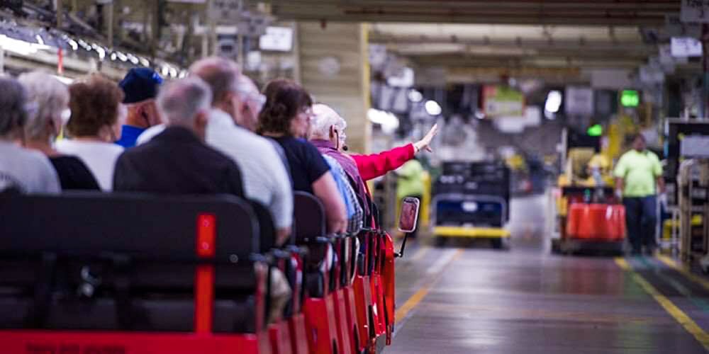 A group of people sit on a tour tram inside a large factory or warehouse; one person is raising a hand while workers move in the background.