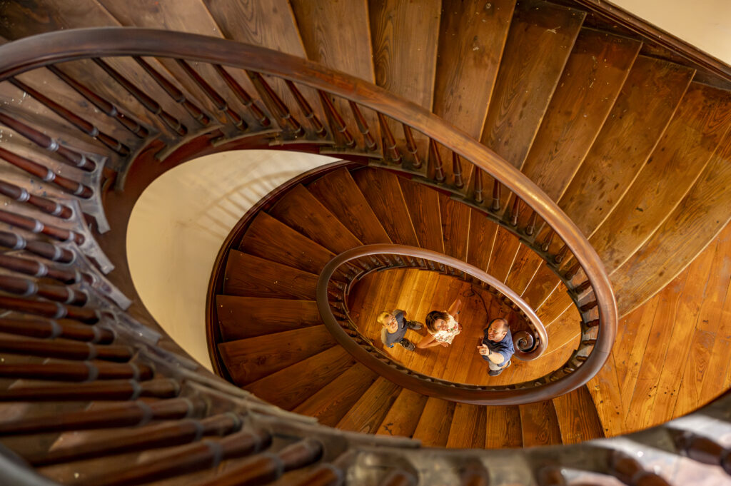 View from above of a spiral wooden staircase with two adults and a child standing at the bottom, looking up. The railing curves tightly around the stairs.