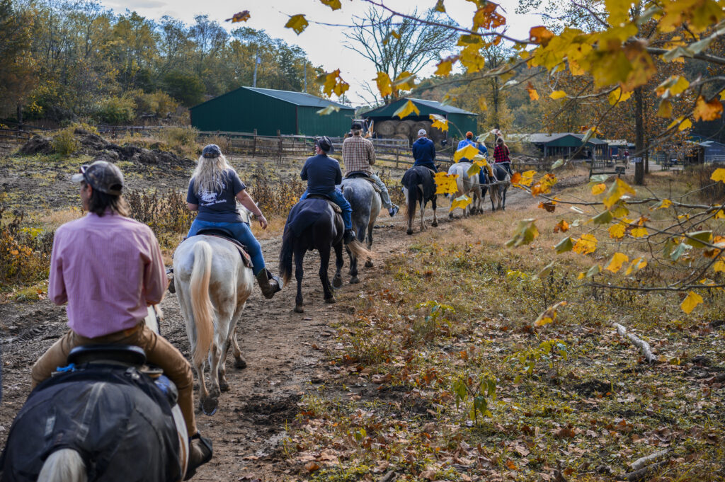 A group of people ride horses along a dirt trail in an autumn landscape, with farm buildings and colorful foliage visible in the background.