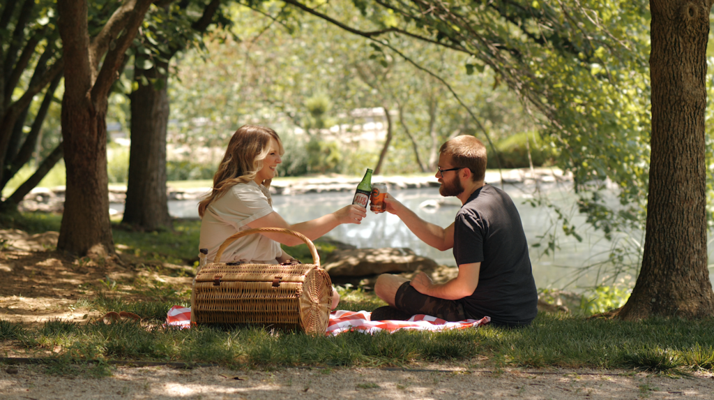 A woman and man sit on a picnic blanket by a lake, clinking bottles. A wicker basket and trees are nearby.