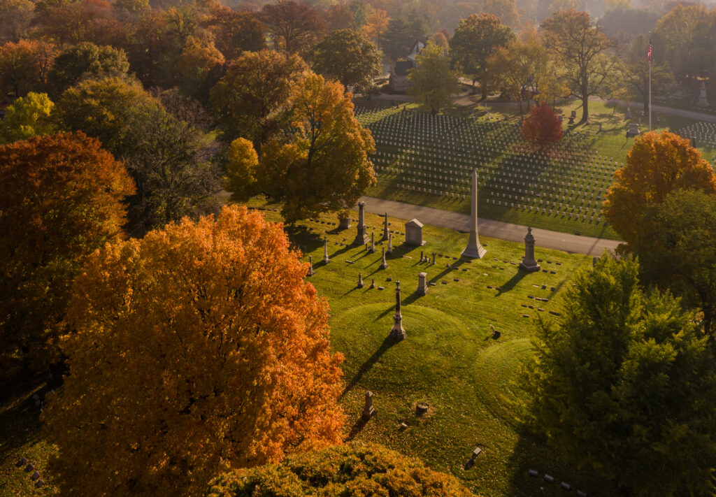 Aerial view of a cemetery in autumn, with rows of gravestones and colorful trees with orange, green, and yellow leaves.
