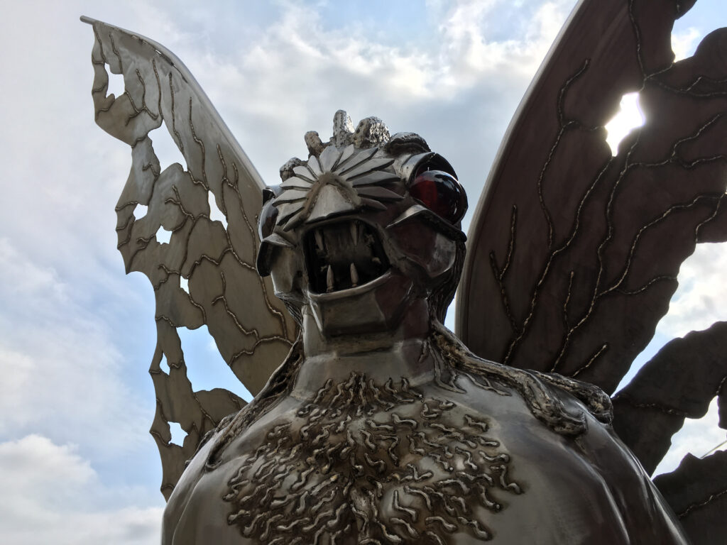 A metallic insect-like statue with large, tattered wings, red eyes, and sharp teeth stands against a cloudy sky.