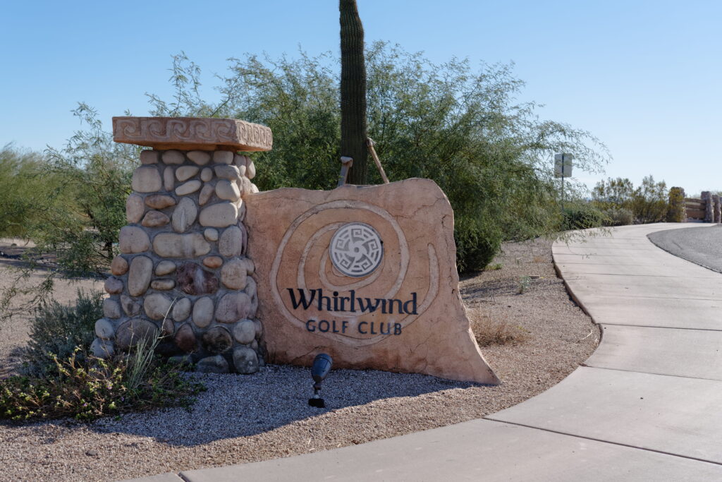 Stone sign for Whirlwind Golf Club next to a rock pillar, surrounded by desert plants, located beside a curved sidewalk and road.