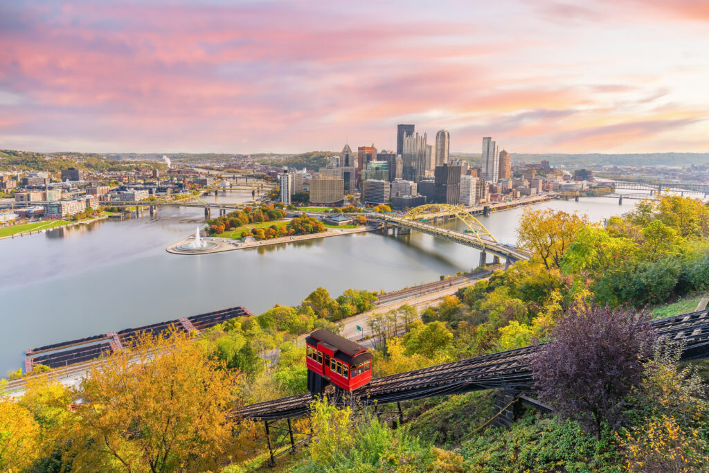 Pittsburgh skyline at sunset with a red incline railway car descending a hill and rivers converging in the foreground.