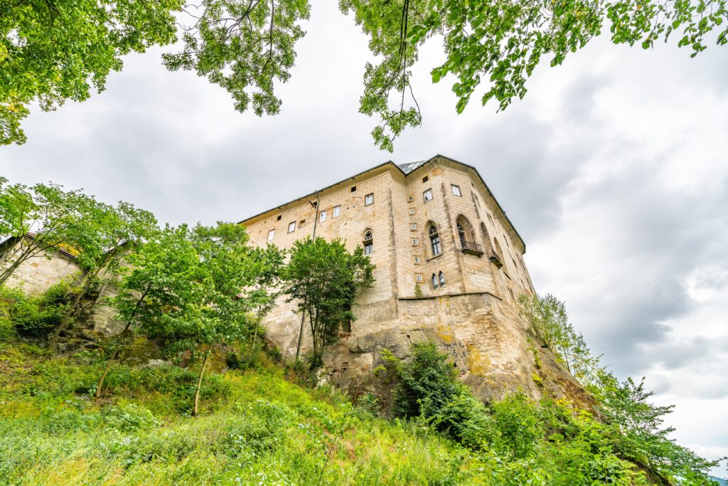 Stone castle perched on a rocky hill, surrounded by green trees and grass under a cloudy sky.
