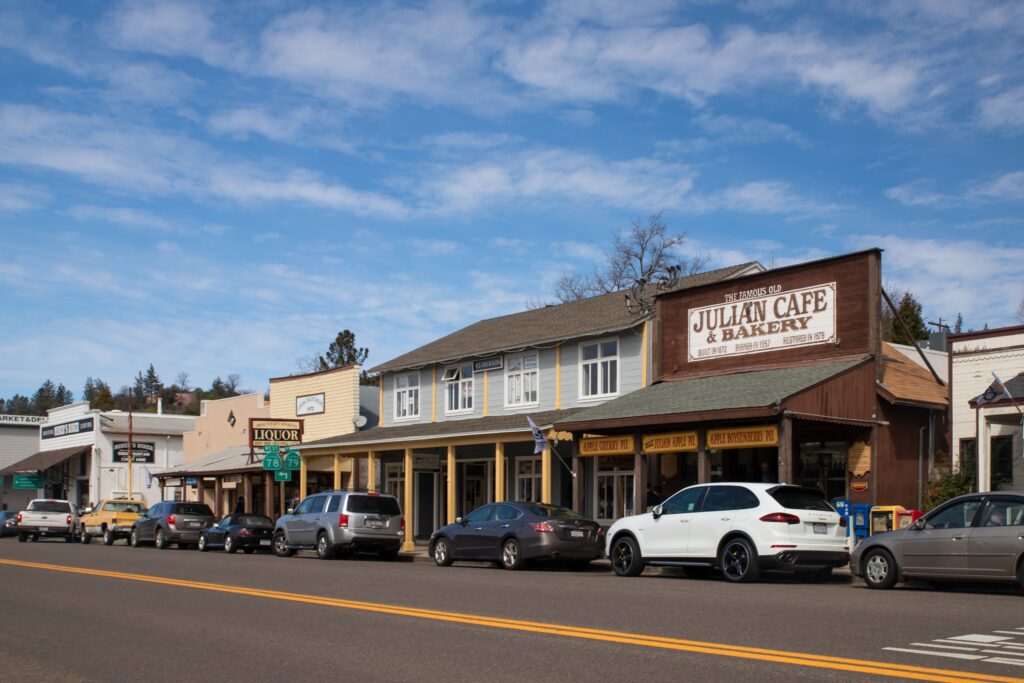 A row of shops, including Julian Cafe & Bakery, line a street with parked cars under a partly cloudy sky in a small town.