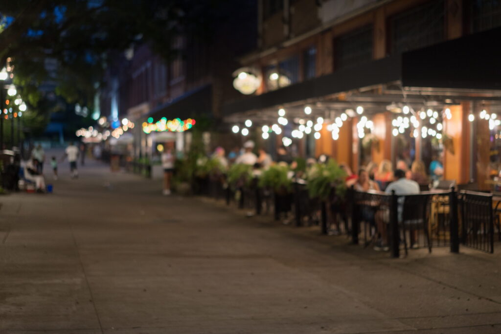 A blurred image of an outdoor restaurant with people seated at tables under string lights along a city sidewalk at night.