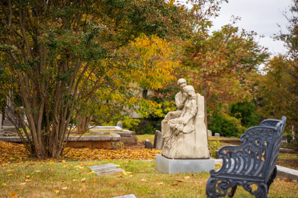Stone statue of a seated figure holding another person in a cemetery, surrounded by autumn trees and fallen leaves; a black metal bench is in the foreground.