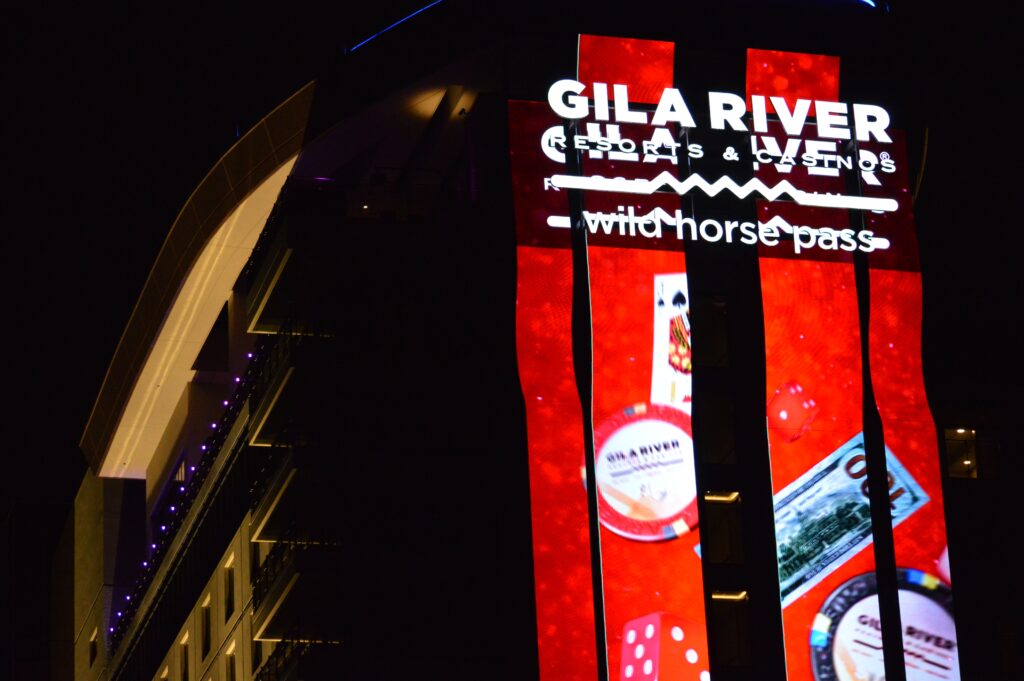 Night view of the Gila River Resorts & Casinos Wild Horse Pass sign, lit up with red graphics featuring casino imagery like cards, chips, and dice.