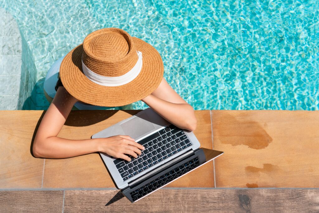 Person wearing a wide-brimmed hat works on a laptop beside a swimming pool, with one arm resting on the pool’s edge.
