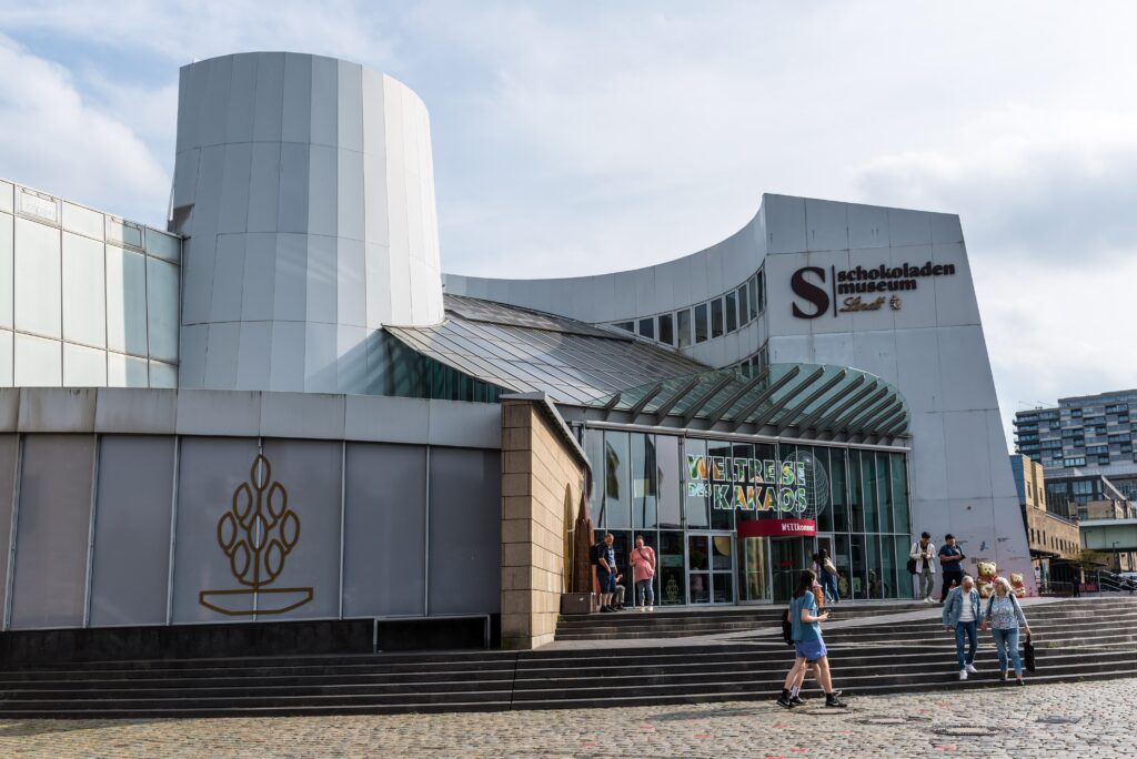 Exterior view of the Schokoladenmuseum in Cologne, Germany, with people standing and walking near the entrance on a cloudy day.