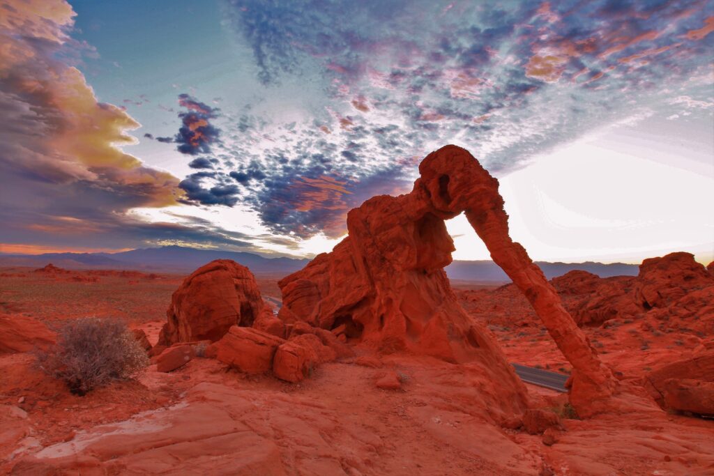 Red sandstone rock formation resembling an elephant stands under a colorful, cloud-filled sky in a desert landscape.