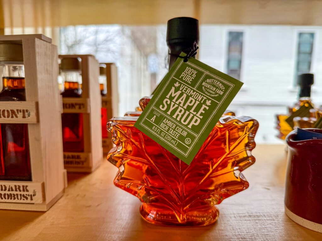 A bottle of Vermont maple syrup in a maple leaf-shaped glass container with a green label sits on a wooden shelf near other syrup bottles.