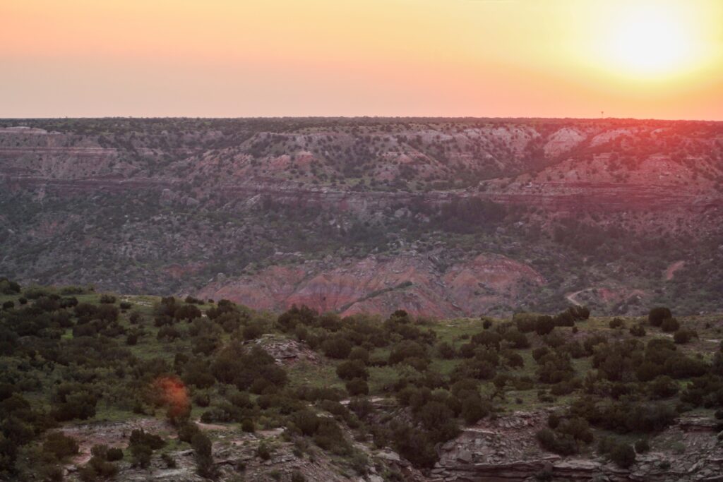 Sunset over a rugged canyon landscape with scattered green bushes and rocky cliffs under an orange sky.