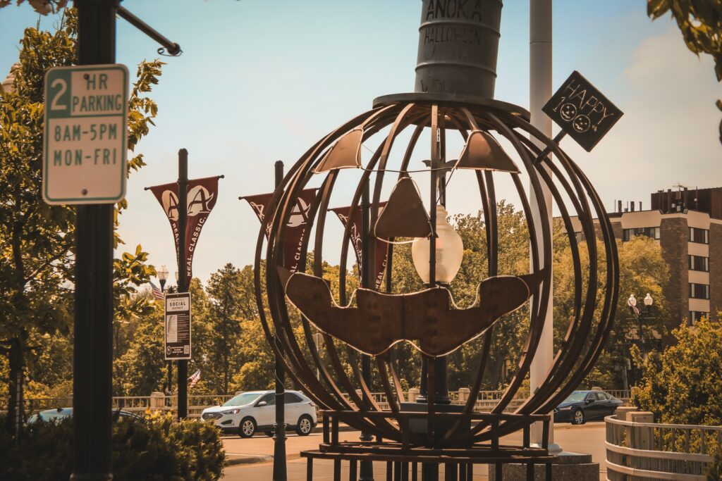 Metal sculpture of a Jack-o’-lantern with a “Happy Halloween” sign, situated near a street and parking sign, surrounded by trees and buildings.