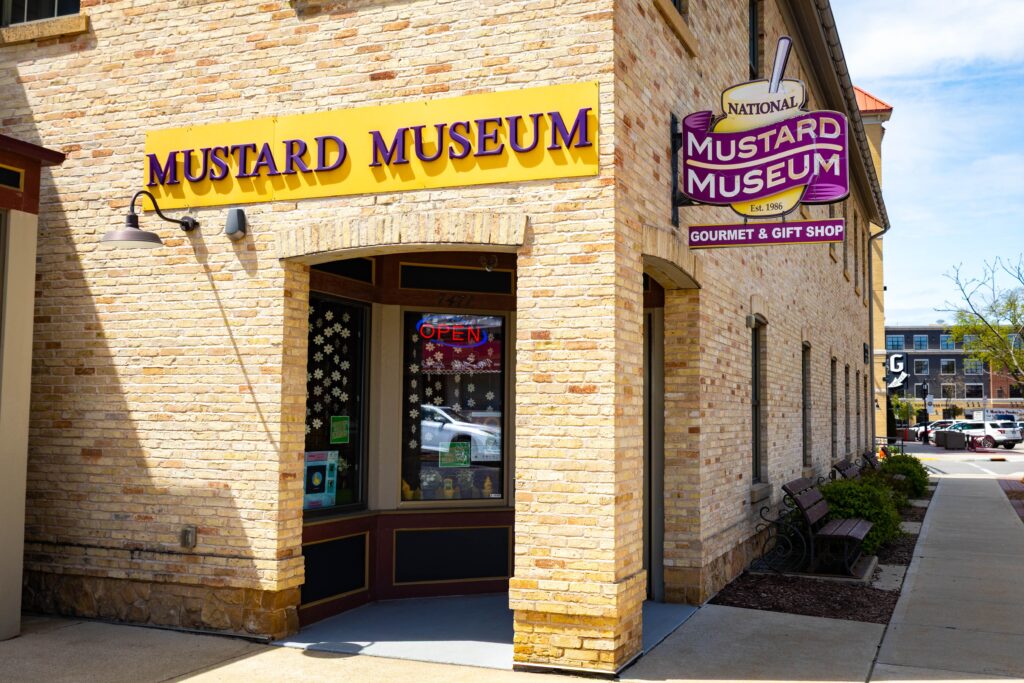 The exterior of the National Mustard Museum, featuring a yellow sign and a corner entrance with an “open” sign in the window.
