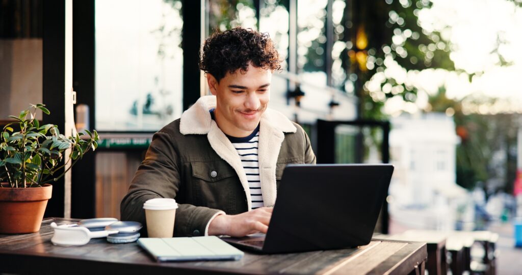 A person sits at an outdoor table, working on a laptop with a coffee cup, plant, and tablet nearby. The setting appears to be a cafe with natural light.