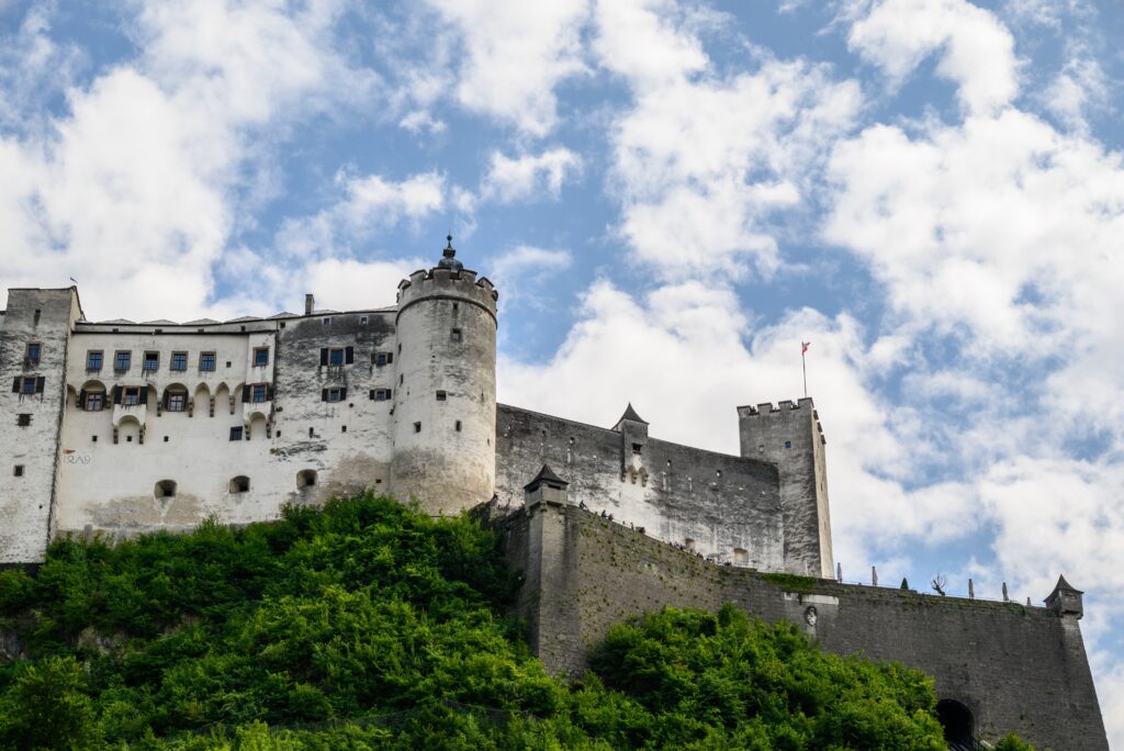 Medieval stone castle with towers and walls sits atop a green hill against a partly cloudy sky.