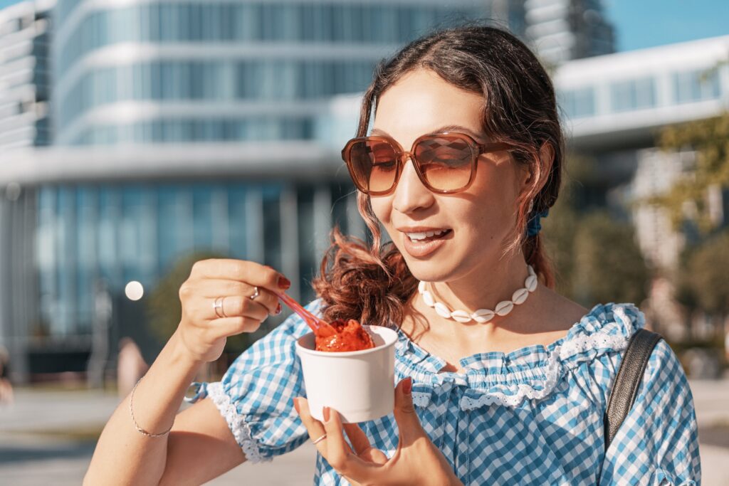A woman wearing sunglasses and a shell necklace holds a cup of red gelato and eats with a spoon outdoors near modern buildings.