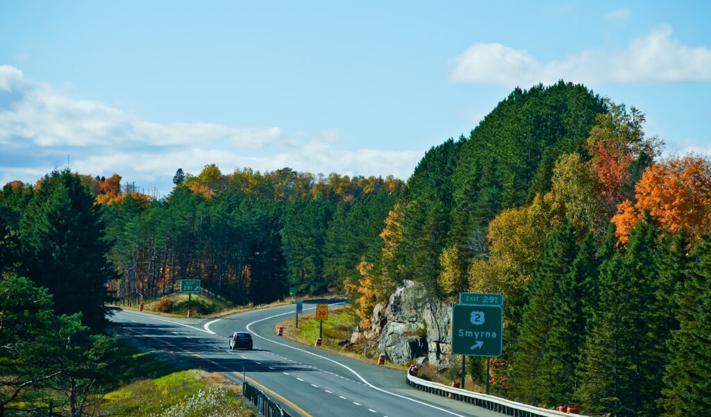 A car drives on a highway surrounded by trees with fall foliage. A road sign indicates Exit 291 for Smyrna. The sky is mostly clear with some clouds.