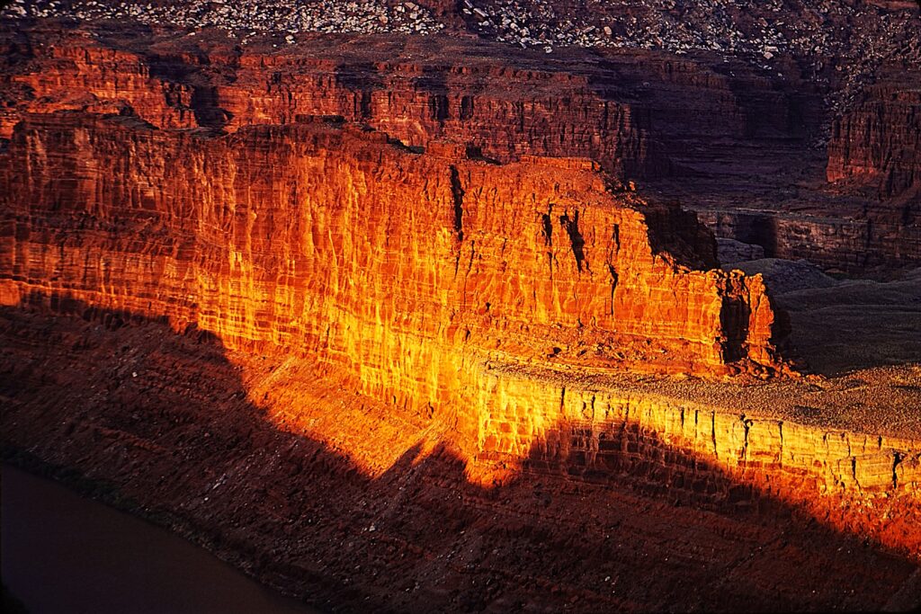 Sunlight illuminates the face of a red rock canyon wall, casting deep shadows; a river runs along the base of the cliffs.