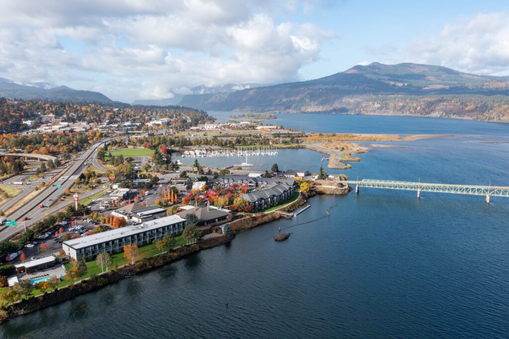 Aerial view of a riverside town with a bridge, marina, highways, and surrounding hills under a partly cloudy sky.