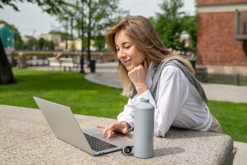 Woman in a white shirt uses a laptop outdoors on a stone table, with a water bottle and smartwatch beside her. Trees and buildings are visible in the background.