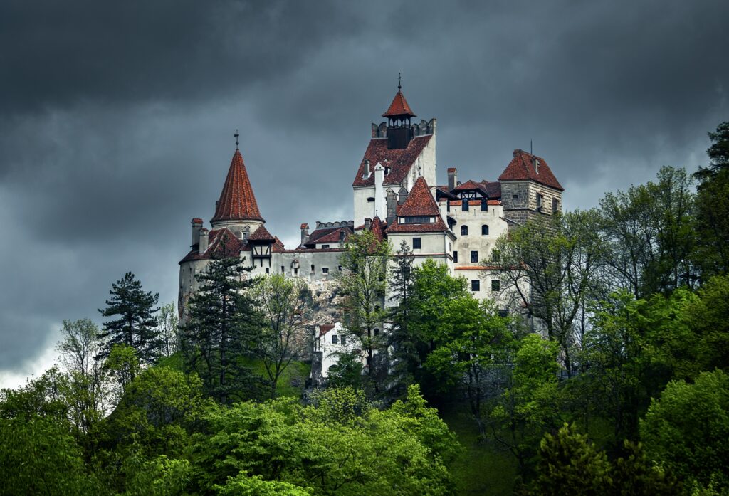 A large medieval castle with red rooftops stands on a forested hill under a cloudy, dark sky.