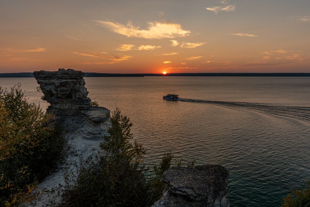 A boat travels across a calm lake at sunset, with rocky cliffs and trees in the foreground and the sun low on the horizon.