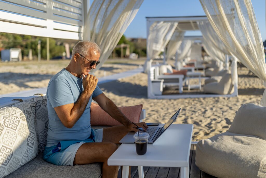 Older man in sunglasses uses a laptop at a shaded beach cabana, with a drink on the table and white curtains blowing in the breeze.