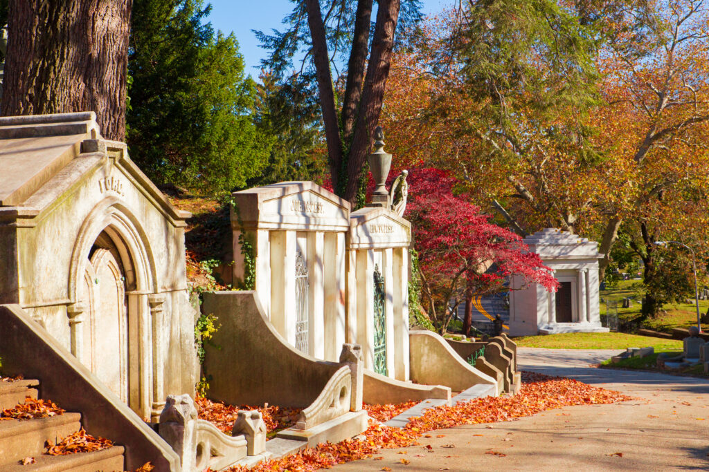 Stone mausoleums and monuments line a cemetery pathway covered with autumn leaves, surrounded by trees with colorful fall foliage in bright daylight.