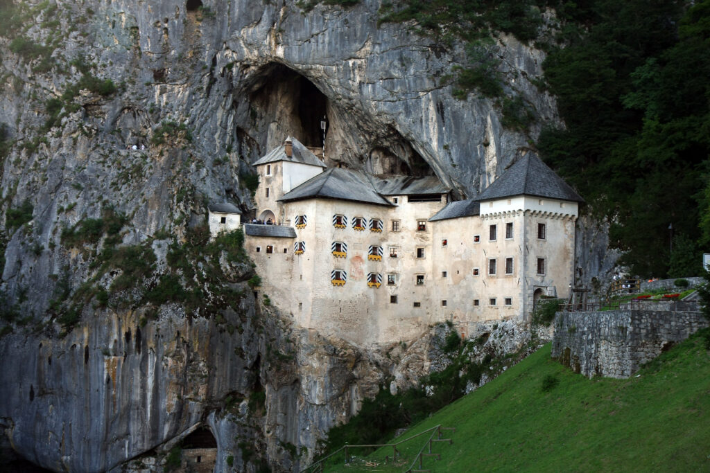 A stone castle is built into the side of a rocky cliff, with several windows and a cave above it, surrounded by greenery.
