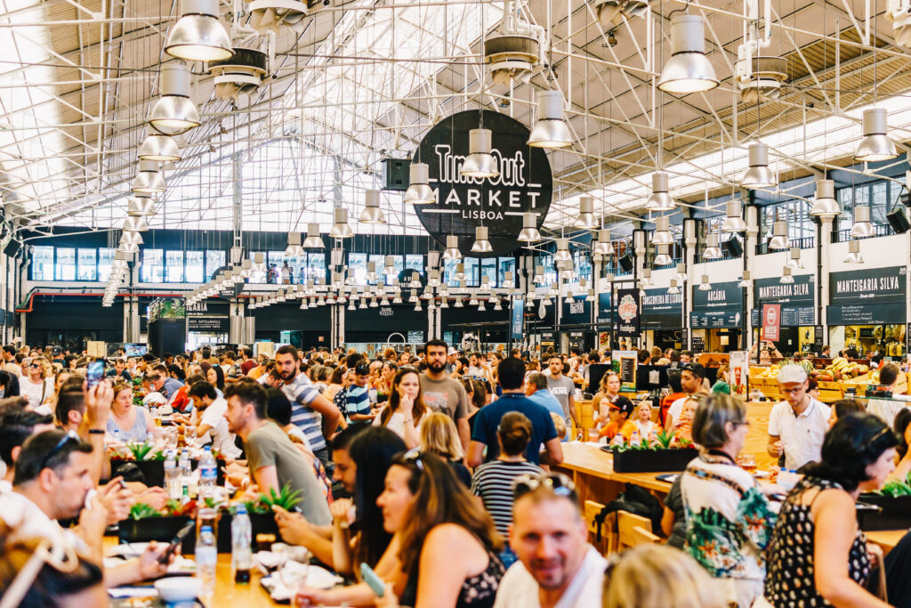 Crowds of people dine and walk inside Time Out Market Lisboa, a large indoor food hall with high ceilings and food vendor stalls lining the sides.
