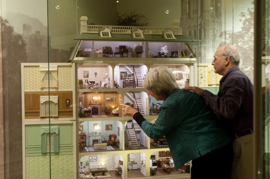 An older couple behind a glass partition looking at and pointing toward the rooms of a large multi-story dollhouse.