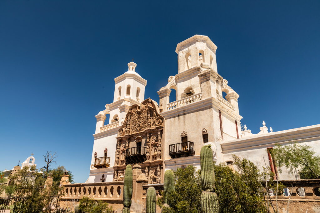 The San Xavier del Bac Mission church with ornate stone facade and two towers under a clear blue sky, surrounded by saguaro cacti.