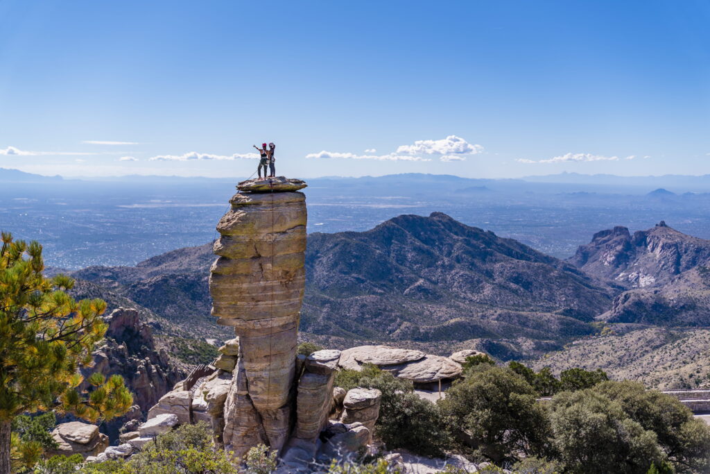 Two hikers stand on top of a tall rock formation overlooking a vast valley.