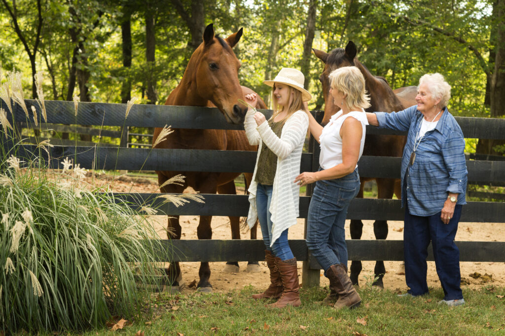 Three generations of women stand by a wooden fence, with the youngest offering a piece of grass to a brown horse.