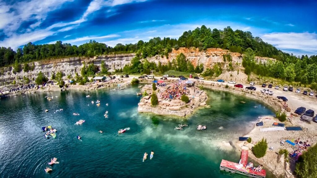 Aerial view of a turquoise rock quarry lake with a central island crowded with people, surrounded by rocky cliffs and cars parked along the perimeter.